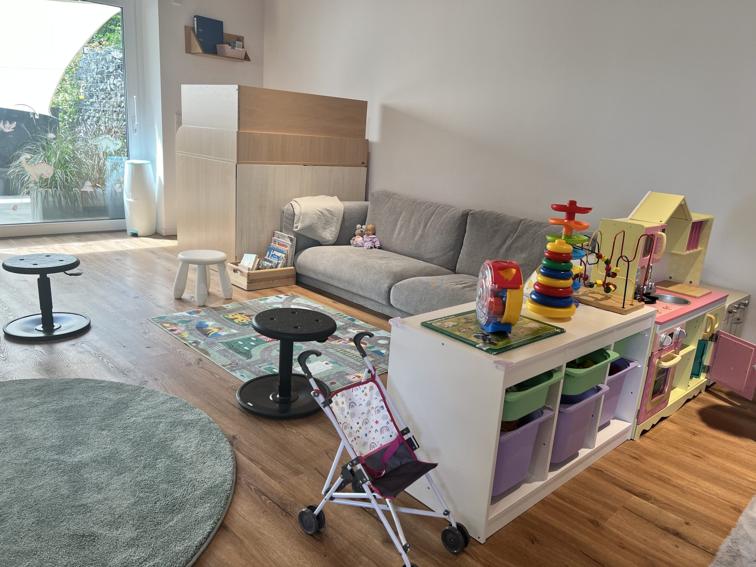 Kinderfreundlicher Raum mit grauem Sofa, Spielteppich, Spielküche und bunten Aufbewahrungsboxen, ideal für kreatives Spielen.
