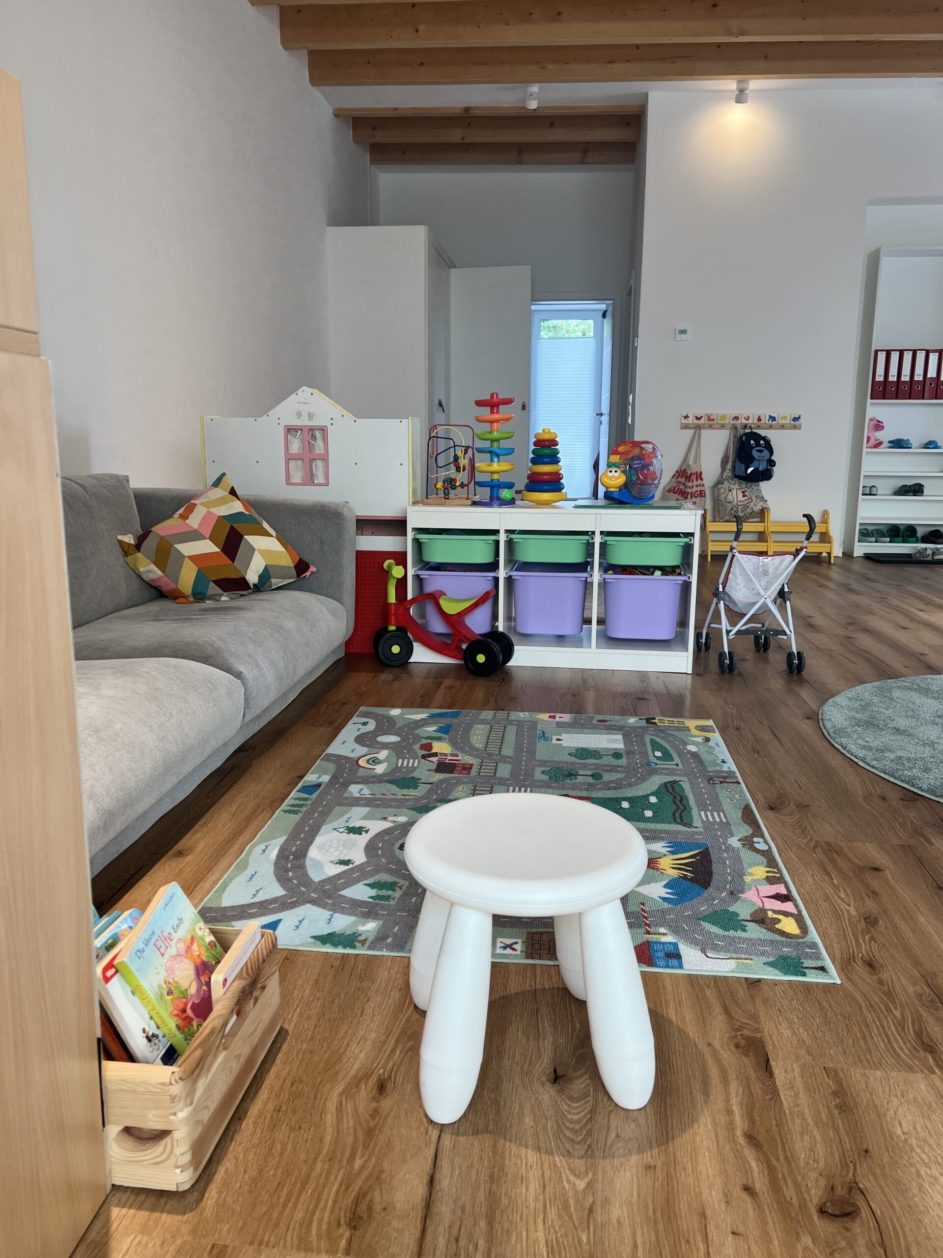 Raum mit Sofa, Spielteppich, niedriger Sitzgelegenheit und Spielregal mit bunten Boxen und Spielküche.
