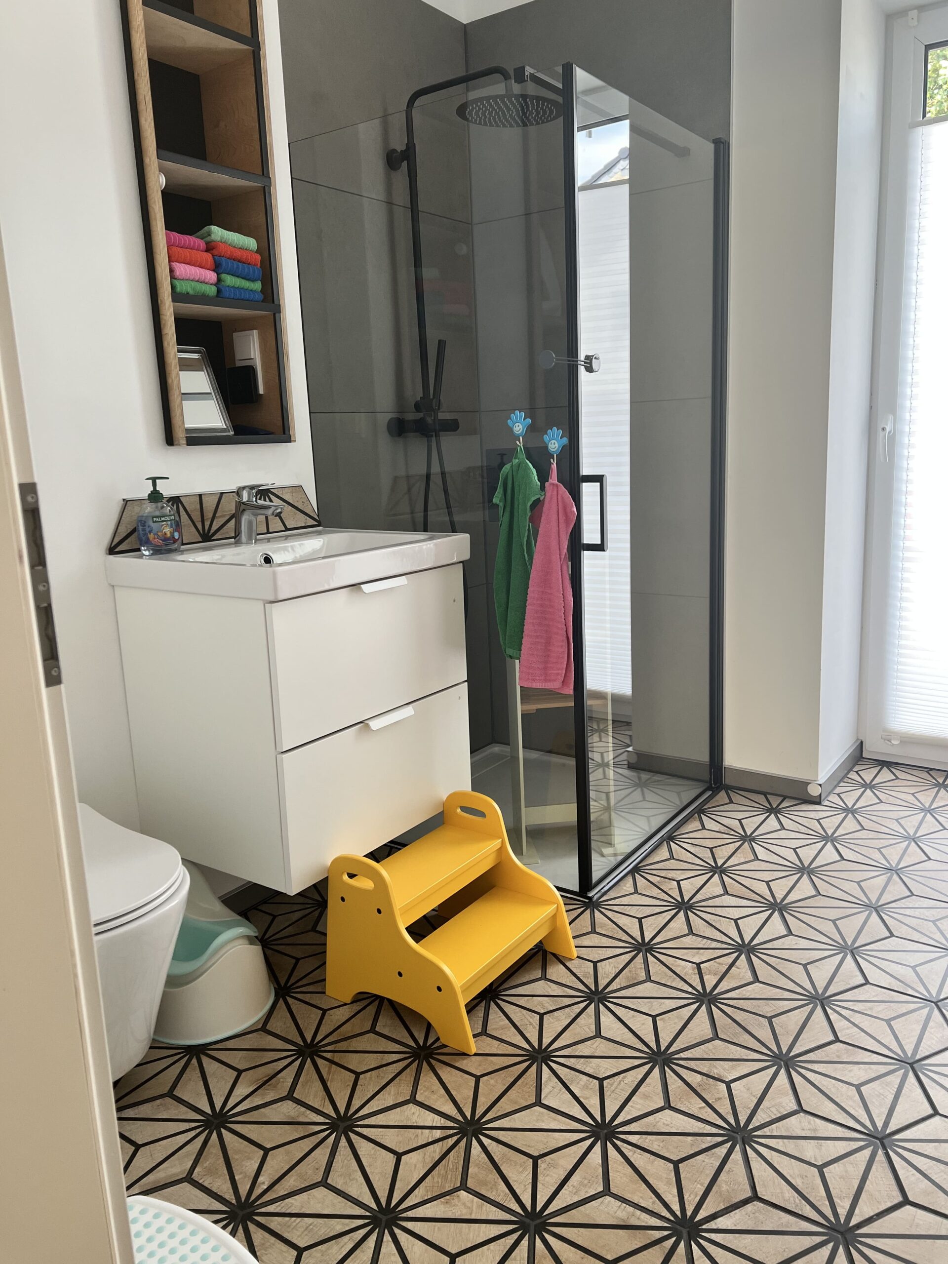 Modernes Bad mit bodentiefer Dusche, farbigen Handtüchern, Waschbecken und gelber Trittstufe für Kinder.